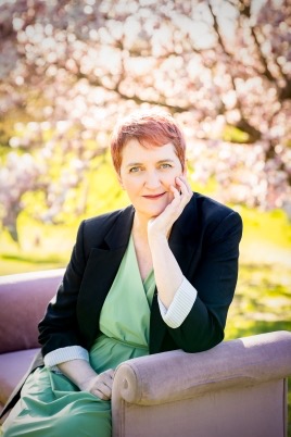 A woman sitting on a chair outdoors, smiling gently while resting her chin on her hand. She has short reddish hair and is wearing a green dress with a black blazer. In the background, there are blossoming trees, creating a soft, colorful atmosphere.