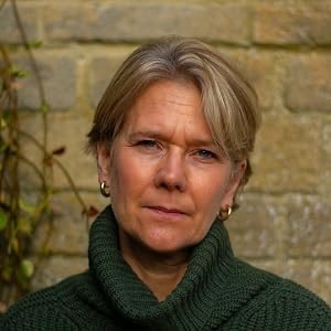A woman with short, light hair wearing a green sweater, standing against a stone wall with greenery.
